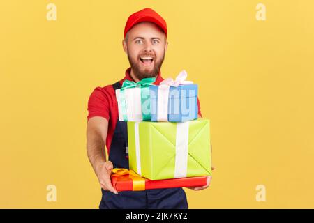 Portrait von aufgeregt erstaunt Lieferung Mann trägt Uniform und rote Kappe geben viele Geschenk-Boxen an Kunden, Tür zu Tür Lieferung, schnellen Service. Innenaufnahme des Studios isoliert auf gelbem Hintergrund. Stockfoto