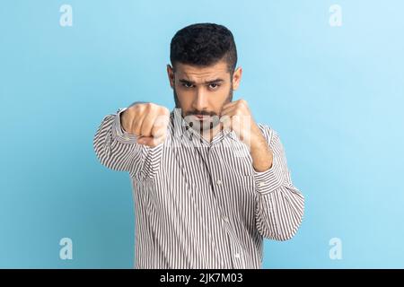 Porträt eines aggressiven jungen erwachsenen Geschäftsmannes, der mit Boxfäusten und bereit zum Angriff oder zur Verteidigung steht, mit einem wütenden Gesicht aussieht und ein gestreiftes Hemd trägt. Innenaufnahme des Studios isoliert auf blauem Hintergrund Stockfoto