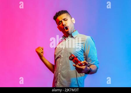 Extrem zufriedener Mann im Hemd, der Videospiel spielt, den Joystick hält, seine siegreiche, geballte Faust feiert und fröhlich schreit. Innenaufnahmen im Studio, isoliert auf farbigem Neonlicht-Hintergrund. Stockfoto