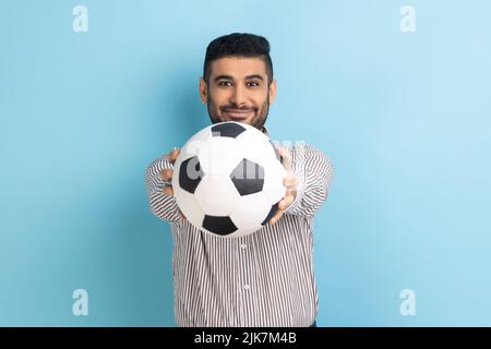 Glücklicher, gutaussehender bärtiger Geschäftsmann, der mit einem lächelnden, positiven Ausdruck den Fußball auf der Hand hält und ein gestreiftes Hemd trägt. Innenaufnahme des Studios isoliert auf blauem Hintergrund. Stockfoto