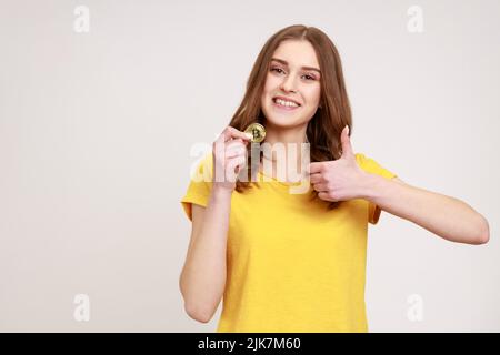 Super Bitcoin. Zufriedene braunhaarige Frau in urbaner Kleidung, die Daumen nach oben zeigt, wie Geste und hält die BTC-Münze, Kryptowährung. Innenaufnahme des Studios isoliert auf grauem Hintergrund. Stockfoto