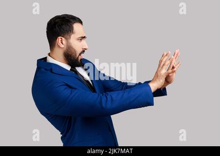 Seitenansicht eines bärtigen Mannes, der die Hände aus plötzlicher Angst hebt, erschrocken aussieht, zu Tode erschrocken ist, einen entsetzlichen Gesichtsausdruck hat, einen offiziellen Anzug trägt. Innenaufnahme des Studios isoliert auf grauem Hintergrund. Stockfoto