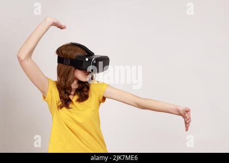 Teenager, die eine Virtual-Reality-Brille am Kopf tragen und Kampfspiel, Cyberspace, VR-Headset-Technologie und gelbes T-Shirt spielen. Innenaufnahme des Studios isoliert auf grauem Hintergrund. Stockfoto