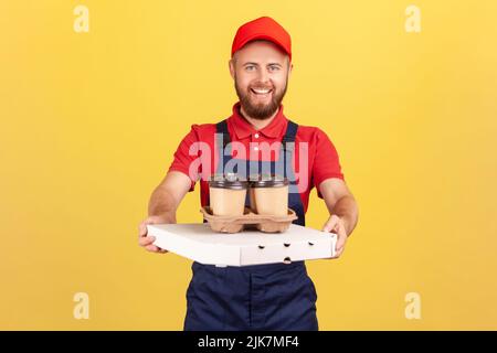 Porträt eines lächelnden professionellen Kuriers mit blauen Overalls, der mit Pizzakarton und Kaffee in einer Einweg-Tasse steht, Fast-Food-Lieferservice. Innenaufnahme des Studios isoliert auf gelbem Hintergrund. Stockfoto