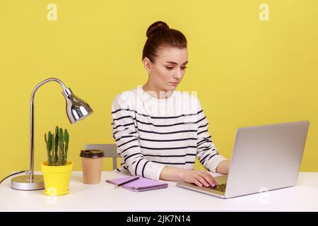 Eine clevere Geschäftsfrau im gestreiften Hemd sitzt am Arbeitsplatz und schaut ernsthaft auf den Laptop-Monitor, macht Notizen, arbeitet an einem Notebook. Studio-Innenaufnahme isoliert auf gelbem Hintergrund. Stockfoto