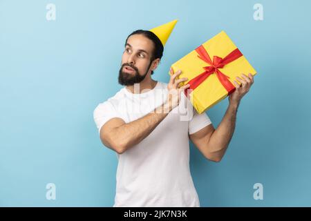 Porträt eines Mannes, der weißes T-Shirt trägt und in einem Partykegel hält und schüttelt, eingewickelte Geschenk-Box, interessiert, was im Inneren, Geburtstag feiert. Innenaufnahme des Studios isoliert auf blauem Hintergrund. Stockfoto