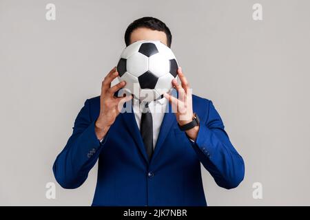 Porträt eines unbekannten anonymen Geschäftsmannes, der seine Lieblingsmannschaft mit Fußballball im offiziellen Anzug unterstützt. Innenaufnahme des Studios isoliert auf grauem Hintergrund. Stockfoto