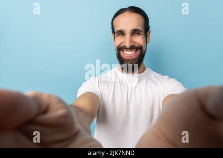 Mann Blogger mit Bart in weißem T-Shirt Blick auf die Kamera mit toothy Lächeln, in guter Laune, Übertragung Livestream, POV, Sicht auf Foto. Innenaufnahme des Studios isoliert auf blauem Hintergrund. Stockfoto