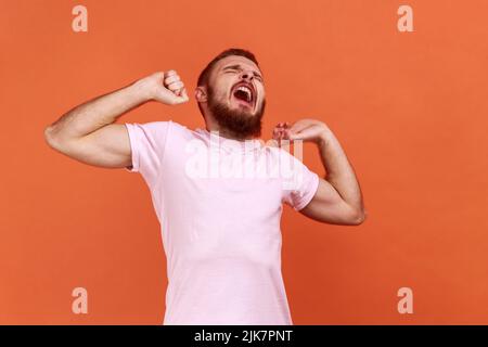 Porträt eines müden bärtigen Mannes, der mit der Hand gähnt und den Mund bedeckt, sich erschöpft fühlt, keinen Schlaf hat, Arme ausstreckt, rosa T-Shirt trägt. Innenaufnahme des Studios isoliert auf orangefarbenem Hintergrund. Stockfoto