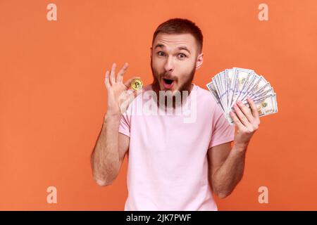 Porträt eines bärtigen Mannes, der Bitcoin- und Dollarnoten hält, die Kamera anschaut, staunende Profite von Kryptowährung in pinkem T-Shirt trägt. Innenaufnahme des Studios isoliert auf orangefarbenem Hintergrund. Stockfoto