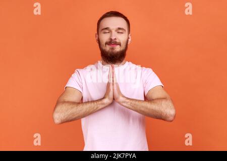 Porträt eines entspannten, gutaussehenden bärtigen Mannes, der in Yoga-Pose steht und versucht zu entspannen, hält die Handflächen zusammen und trägt ein rosa T-Shirt. Innenaufnahme des Studios isoliert auf orangefarbenem Hintergrund. Stockfoto