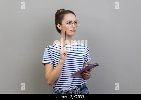Porträt einer Frau, die gestreiftes T-Shirt trägt und Notizen in einem Notizbuch macht, Geschäftsidee schreibt, Zukunftspläne plant und mit durchdachtem Ausdruck wegschaut. Innenaufnahme des Studios isoliert auf grauem Hintergrund. Stockfoto