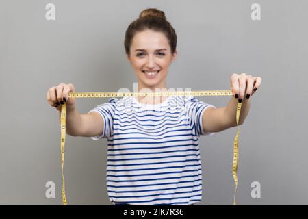 Porträt einer fröhlichen, positiven Designerin oder Näherin in gestreiftem T-Shirt mit Maßband, Schneiderin, die arbeitet, misst. Innenaufnahme des Studios isoliert auf grauem Hintergrund. Stockfoto