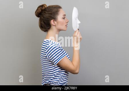 Seitenansicht Porträt einer Frau, die gestreiftes T-Shirt trägt, die weiße Maske mit aufmerksamem Blick hält und versucht, die verborgenen Persönlichkeit zu verstehen. Innenaufnahme des Studios isoliert auf grauem Hintergrund. Stockfoto