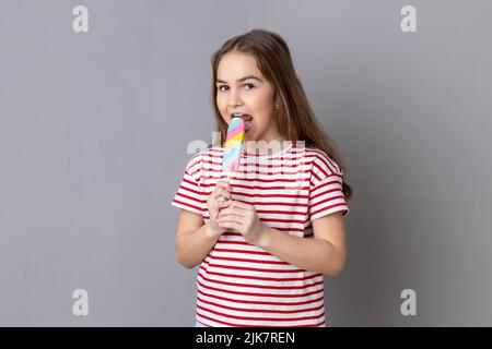 Portrait von entzückten niedlichen kleinen Mädchen trägt gestreifte T-Shirt beißen bunten Süßigkeiten, mit kindlichem zufriedenen Ausdruck wegschauen. Innenaufnahme des Studios isoliert auf grauem Hintergrund. Stockfoto