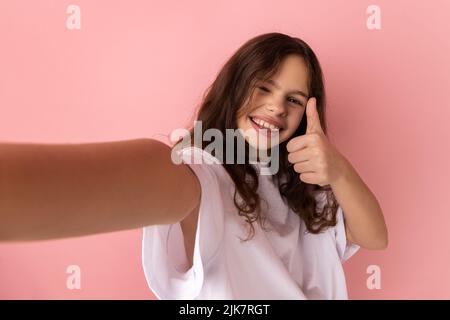 Kleines Mädchen trägt weißes T-Shirt hat optimistischen Gesichtsausdruck, macht Selfie, drückt aufrichtige Emotionen, zeigt Daumen nach oben, Standpunkt Foto. Innenaufnahme des Studios isoliert auf rosa Hintergrund. Stockfoto