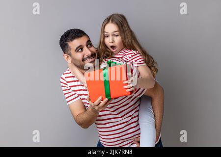 Porträt eines glücklichen Vaters, der seine überraschte Tochter auf dem Rücken hält und ihr zum Geburtstag ein Geschenk macht, Familie trägt gestreifte T-Shirts. Innenaufnahme des Studios isoliert auf grauem Hintergrund. Stockfoto