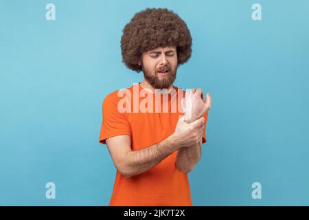 Porträt eines Mannes mit Afro-Frisur im T-Shirt, massiert Wunden Arm, Gefühl taub steife Muskeln, Symptom des Karpaltunnelsyndroms, Gelenkentzündung. Innenaufnahme des Studios isoliert auf blauem Hintergrund. Stockfoto