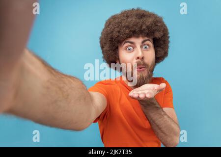 Porträt eines gutaussehenden Mannes mit Afro-Frisur, der ein orangefarbenes T-Shirt trägt und Luftküsse an seine Abonnenten sendet, mit Posen und Lippen posiert, Sichtbild. Innenaufnahme des Studios isoliert auf blauem Hintergrund. Stockfoto