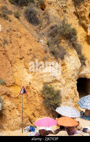 Lagos, Portugal. 21.. Juli 2022. Urlauber liegen und sitzen in der Mittagshitze unter Sonnenschirmen am Strand 'Praia do Camilo' neben einem Felsen mit einem Schild, das vor fallenden Steinen warnt. Quelle: Viola Lopes/dpa/Alamy Live News Stockfoto