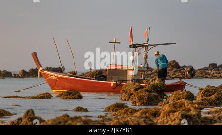 Ein thailändisches Longtail-Boot, das zwischen Felsen am Strand festgemacht wurde und der Kapitän seine Angelausrüstung vorbereitete Stockfoto