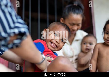 Ein junger thailändischer Junge lächelt, als er von seinem Sponsor gekitzelt wird, während seine Mutter und seine Schwestern hinter dem verschwommenen Hintergrund sitzen Stockfoto