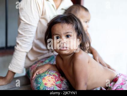 Ein junges Thai-Mädchen mit schmutzigem Gesicht starrt auf die Kamera, während sie sich auf den Schoß ihrer Mutter lehnt Stockfoto