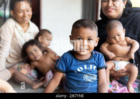 Ein junger Thai-Junge blickt auf die Kamera, und seine lächelnde Familie sitzt dahinter. Bruder, Schwestern, Mutter, Freund. Thailand, arm, ländlich. Stockfoto