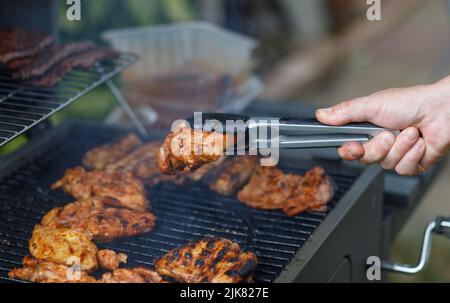 Männliche Hand hält Zange mit Grill Huhn auf einem Metall-grill im Freien im Sommer. Nahaufnahme der Hand, des Handgelenks Stockfoto