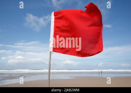 Rote Flagge am Strand. An einem windigen, aber sonnigen Sommertag weht eine rote Flagge im Wind, die Schwimmer vor gefährlichen Surfbedingungen warnt. Rote Flagge, Stockfoto