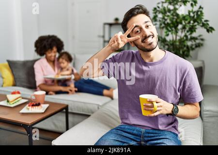 Hispanischer Vater einer interrassischen Familie, der eine Tasse Kaffee trinkt und dabei das Friedenssymbol mit den Fingern über dem Gesicht macht, lächelt fröhlich und zeigt den Sieg Stockfoto