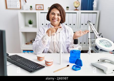 lateinamerikanische Zahnärztin im mittleren Alter, die in der medizinischen Klinik arbeitet, staunt und lächelt zur Kamera, während sie mit der Hand zeigt und mit dem Finger zeigt. Stockfoto