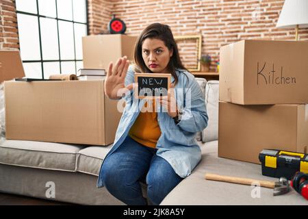 Junge hispanische Frau hält eine Tafel mit einem neuen Heimtext mit offener Hand und tut Stoppschild mit ernstem und selbstbewusster Ausdruck, Verteidigungsgeste Stockfoto