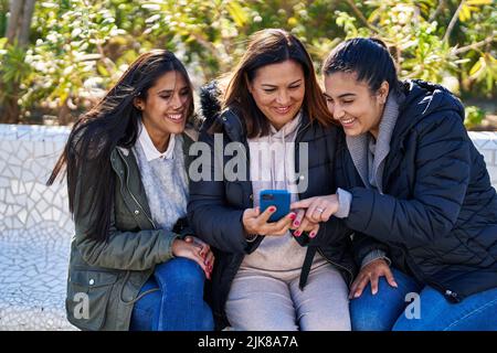 Drei weibliche Mutter und Töchter mit Smartphone sitzen auf der Bank im Park Stockfoto