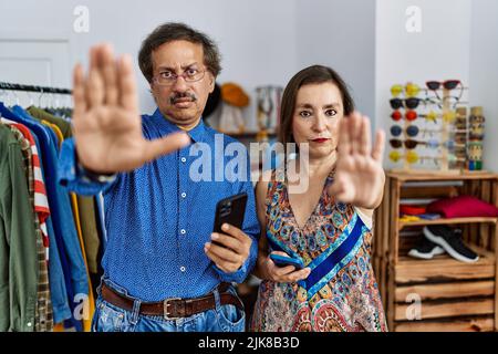 Interracial Paar im mittleren Alter im Einzelhandel mit Smartphone tun aufhören zu singen mit Handfläche der Hand. Warnausdruck mit negativen und schweren gest Stockfoto