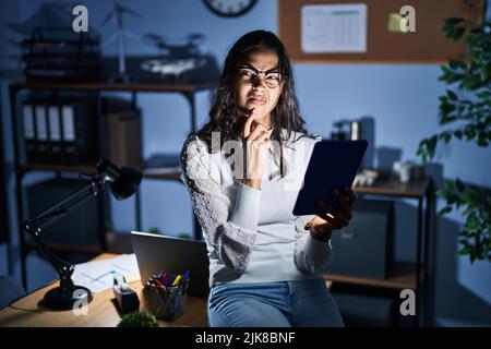 Die junge brasilianerin, die nachts das Touchpad benutzt, arbeitet im Büro und denkt besorgt über eine Frage, besorgt und nervös mit der Hand am Kinn Stockfoto