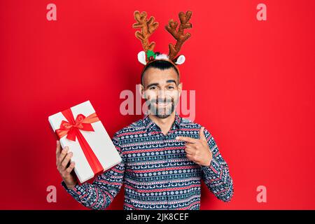 Junger hispanischer Mann trägt Hirsch weihnachtshut mit Geschenk lächelnd glücklich zeigt mit Hand und Finger Stockfoto