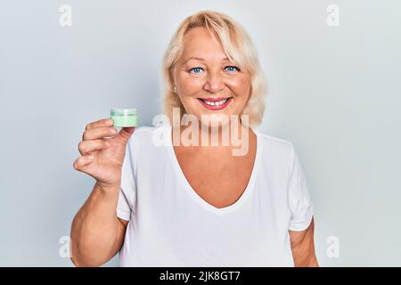 Mitteljährige blonde Frau hält kosmetische Feuchtigkeitscreme Gesichtscreme positiv und glücklich stehend und lächelnd mit einem selbstbewussten Lächeln zeigt Zähne Stockfoto