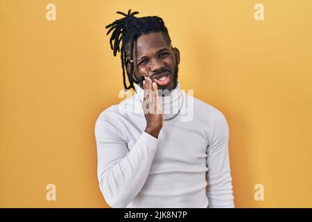 Afrikanischer Mann mit Dreadlocks trägt Rollkragenpullover über gelbem Hintergrund berühren Mund mit Hand mit schmerzhaftem Ausdruck wegen Zahnschmerzen oder Stockfoto