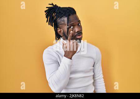 Afrikanischer Mann mit Dreadlocks trägt Rollkragenpullover über gelbem Hintergrund zeigt auf das Auge beobachten Sie Geste, verdächtiger Ausdruck Stockfoto