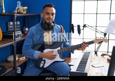 Junger hispanischer Musiker, der mit einem Lächeln auf den Lippen lächelt und im Musikstudio elektrische Gitarre spielt Stockfoto