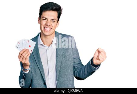 Junger hispanischer Geschäftsmann, der Poker spielt und stolz seine Karten hält, feiert Sieg und Erfolg mit erhobenem Arm Stockfoto