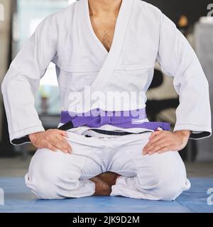 Dein bely hebt deine Fortschritte hervor. Ein nicht erkennbarer junger Mann kniet im vollen Jiu Jitsu gi im Fitnessstudio. Stockfoto