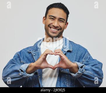 Viel Liebe. Beschnittenes Porträt eines hübschen jungen Mannes, der im Studio vor grauem Hintergrund eine Herzgeste macht. Stockfoto