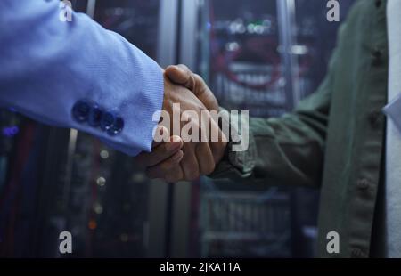 Zwei nicht erkennbare männliche IT-Support-Mitarbeiter schütteln sich in einem dunklen Netzwerk-Serverraum die Hände. Stockfoto