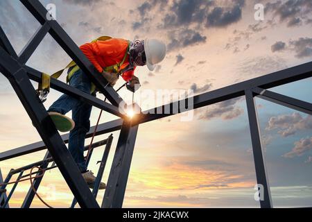 Ein Schweißer schweißen Stahl auf einem Stahldachstuhl. Arbeiten in der Höhe der Ausrüstung. Fallschutzvorrichtung für Arbeiter mit Haken für den Sicherheitsgurt. Arbeiten Stockfoto