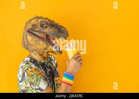 Mann in Dinosaurier Tiermaske trinken Cocktail trägt LGBTQ Stolz Regenbogen Armband, isoliert auf orangefarbenem Hintergrund Stockfoto