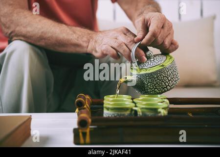 Hände eines älteren Mannes, der Tee aus der Teekannen in Tassen auf dem Bambusblech gießt Stockfoto