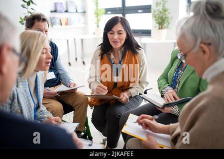 Aufgeregte ältere Menschen, die an einer Gruppentherapie im Pflegeheim teilnehmen, positive ältere Männer und Frauen, die im Kreis sitzen und mit ihnen sprechen Stockfoto