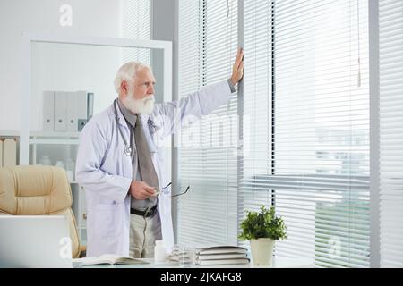 Nachdenklicher Oberarzt, der in seinem Büro am Fenster steht Stockfoto